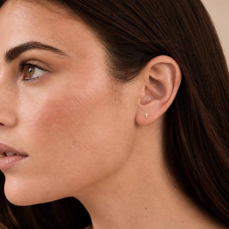 May include: A small, minimalist gold cross stud earring worn in the earlobe. The earring features a simple, polished metal cross design with a post back, providing a subtle and understated look for everyday wear in a single ear piercing.