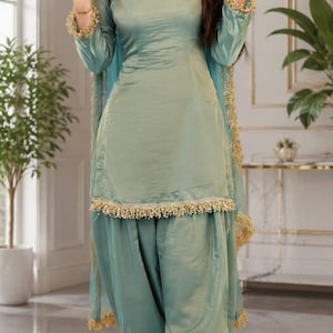 Op de afbeelding: Een saliegroene salwar kameez-set bestaande uit een tuniek met lange mouwen, een vierkante halslijn en een bijpassende geplooide broek. Het ensemble is afgewerkt met gouden franjes langs de zoom, de manchetten en een bijpassende transparante dupatta-sjaal.