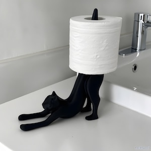 May include: A black cat-shaped toilet roll holder. The holder is made of a solid black material and features a cat stretching with its front paws extended. A white roll of toilet paper is held in place by the cat's back and a black post.