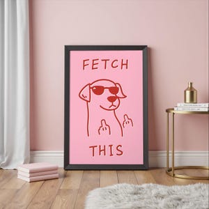 May include: Framed pink art print featuring a cartoon dog wearing sunglasses and giving the middle finger. The words "FETCH THIS" are printed in red above and below the dog illustration. The frame is black.