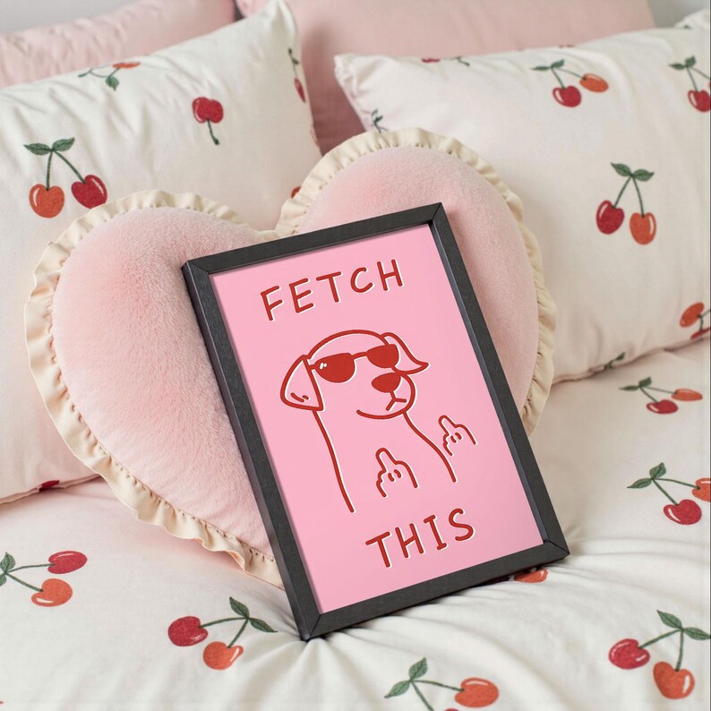 May include: A framed pink print with the text "FETCH THIS" and a cartoon dog wearing sunglasses. The print is set against a pink heart-shaped pillow and cherry-patterned bedding.