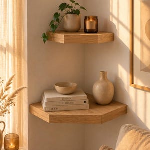 Op de afbeelding: Twee lichtgekleurde houten hoekplanken gemonteerd aan een muur. De bovenste plank bevat een kleine potplant en een glazen kaarsenpot. De onderste plank bevat een stapel van drie boeken met de titels Kinfolk, This is Home en at 100, een kom en een vaas.