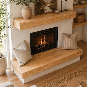 Op de afbeelding: Een gezellige woonkamer met een open haard, houten mantel en haard. Een landschapsschilderij hangt boven de open haard en twee kussens liggen op de haard. Een vaas met groen en kaarsen.