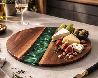 May include: A circular wooden serving board with a green resin inlay, laden with cheese, grapes, olives, and figs. A cheese knife is placed beside it. The board sits on a light grey surface, with wine and other food items in the background.