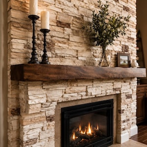 May include: A fireplace with a stacked stone surround in shades of tan, cream, and brown. A dark brown wooden mantel sits above a black metal fireplace insert with visible flames. Two white pillar candles and a glass vase with greenery are present.