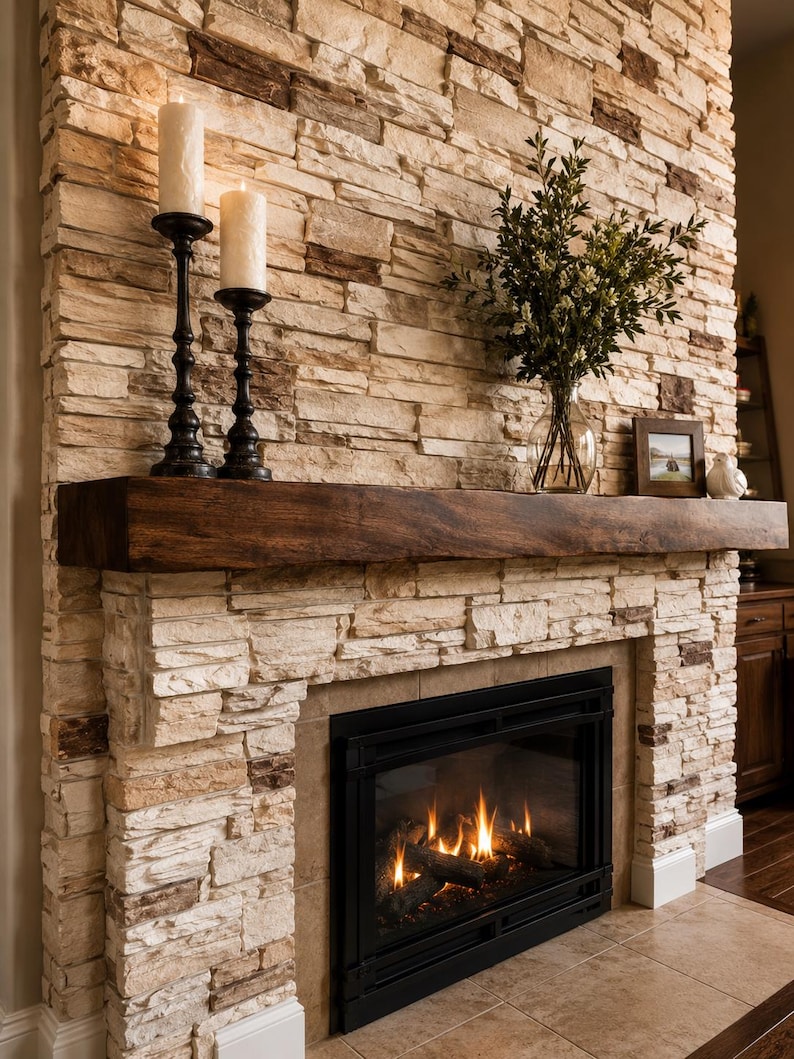 May include: A fireplace with a stacked stone surround in shades of tan, cream, and brown. A dark brown wooden mantel sits above a black metal fireplace insert with visible flames. Two white pillar candles and a glass vase with greenery are present.