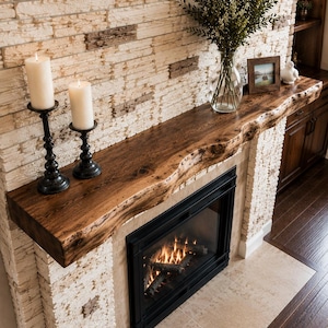 May include: A live edge wooden fireplace mantel mounted above a black framed fireplace insert. The mantel features a natural dark wood grain and irregular front edge, topped with two white pillar candles in black holders and a glass vase.