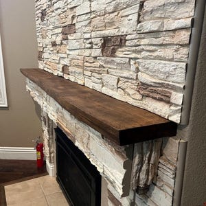 May include: A dark brown wooden fireplace mantel shelf mounted horizontally against a wall covered in stacked, textured stone tiles in shades of cream, beige, and brown. The mantel features a smooth, flat top surface and a rectangular profile.