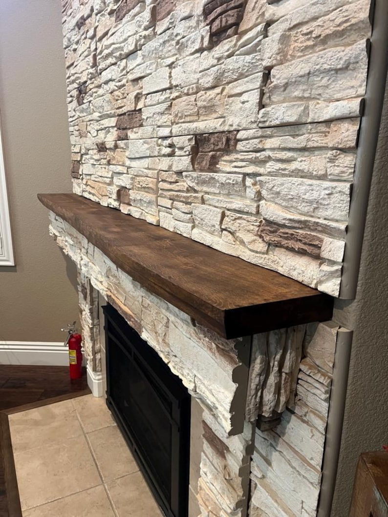 May include: A dark brown wooden fireplace mantel shelf mounted horizontally against a wall covered in stacked, textured stone tiles in shades of cream, beige, and brown. The mantel features a smooth, flat top surface and a rectangular profile.