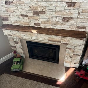 May include: A fireplace featuring a dark brown wooden mantelpiece and a surround constructed from light beige and tan stacked stone tiles. The fireplace insert is black with a glass front, positioned above a square beige tile hearth floor.