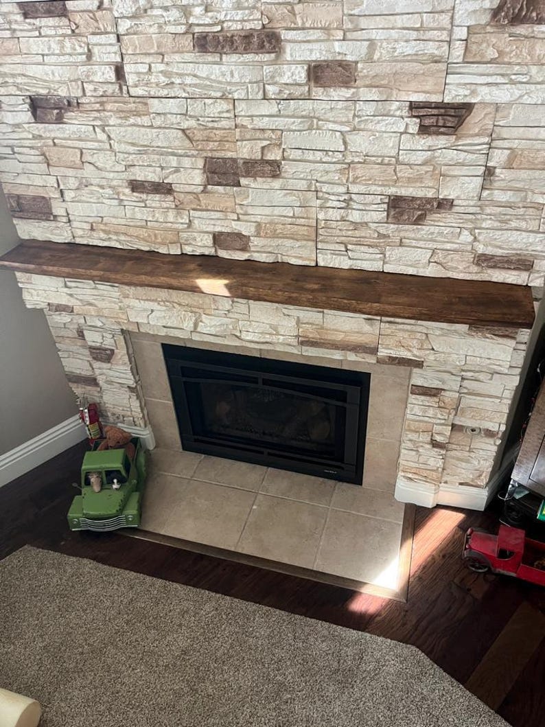 May include: A fireplace featuring a dark brown wooden mantelpiece and a surround constructed from light beige and tan stacked stone tiles. The fireplace insert is black with a glass front, positioned above a square beige tile hearth floor.
