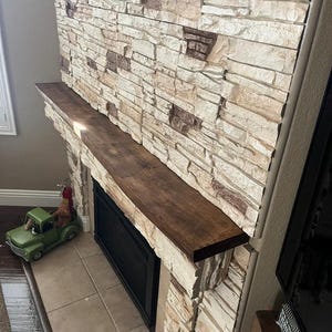 May include: A fireplace mantel made of dark brown wood installed above a black fireplace insert. The wall surrounding the fireplace is covered in stacked stone veneer featuring a mix of cream, beige, and dark brown rectangular stone pieces.