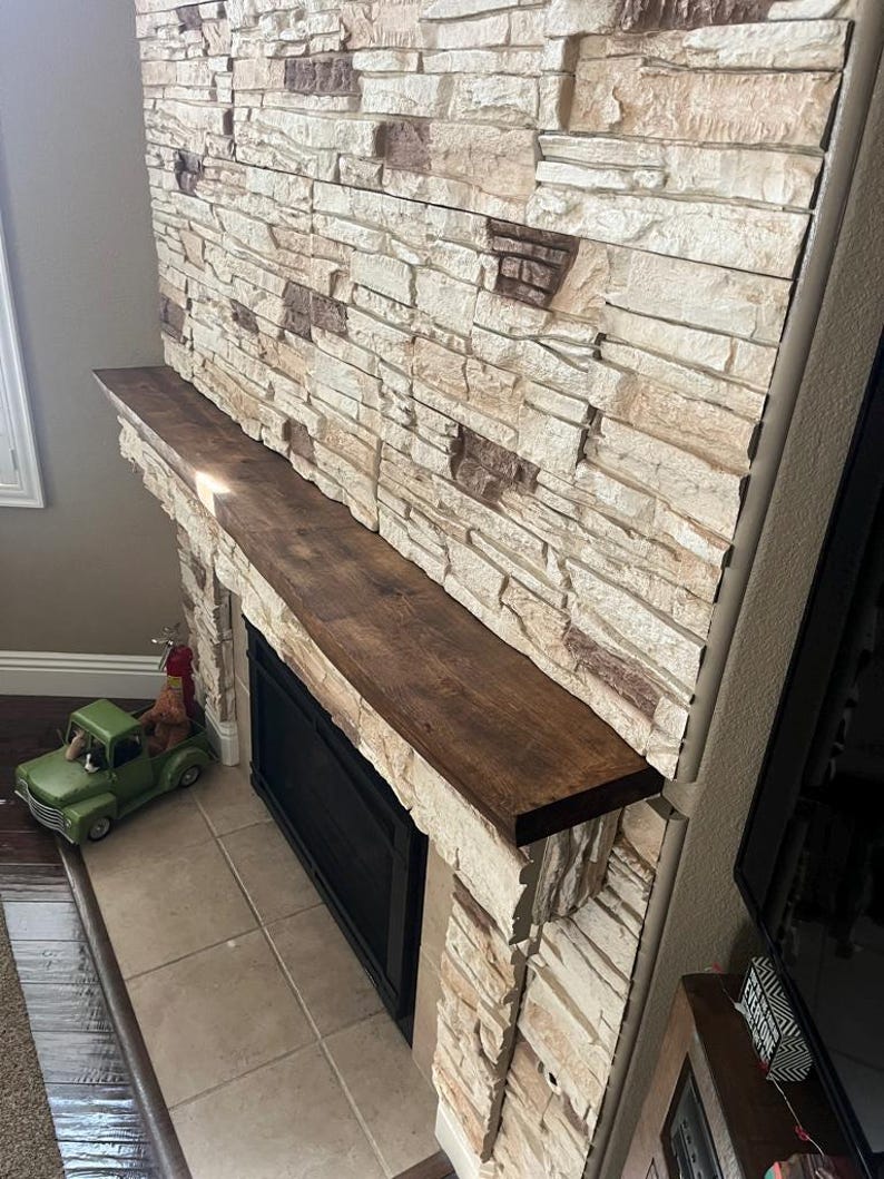 May include: A fireplace mantel made of dark brown wood installed above a black fireplace insert. The wall surrounding the fireplace is covered in stacked stone veneer featuring a mix of cream, beige, and dark brown rectangular stone pieces.