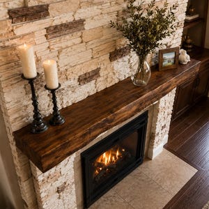 May include: A fireplace with a dark wood mantel, a black metal firebox with visible flames, and a surround made of stacked light-colored stone. Two tall white pillar candles in black holders, a glass vase with greenery, and a small framed picture.