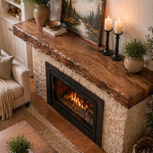 May include: A fireplace with a rough-hewn dark wood mantel, a light-colored stone surround, and a black metal firebox. The mantel holds a landscape painting of a forest, two white pillar candles in black holders, and a ceramic vase with greenery.
