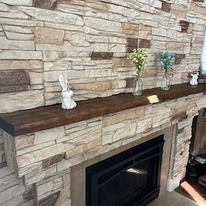 May include: A dark brown wooden fireplace mantel featuring a white ceramic rabbit figurine wearing black glasses, two clear glass bud vases containing yellow and blue flowers, and a small white animal figurine on the far right side.