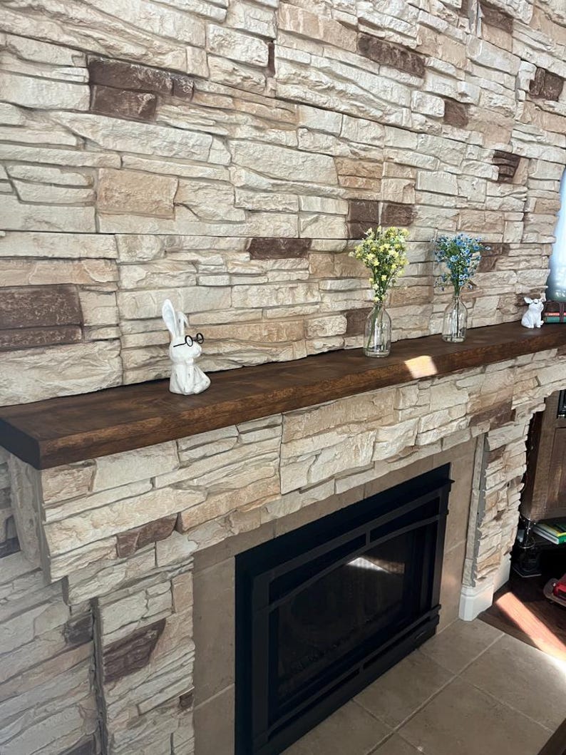 May include: A dark brown wooden fireplace mantel featuring a white ceramic rabbit figurine wearing black glasses, two clear glass bud vases containing yellow and blue flowers, and a small white animal figurine on the far right side.