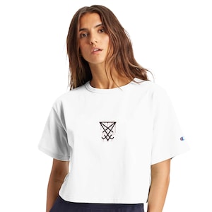 Op de afbeelding: Witte crop top met een zwart en rood grafisch ontwerp. De shirt heeft korte mouwen en een klein Champion-logo op de linkermouw. De afbeelding is een gestileerd symbool.