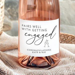 May include: A bottle of rosé champagne with a label that reads "Pairs Well With Getting Engaged." The label also includes the names "Kate & Ray" and two champagne glasses. The bottle is on a woven tray with a basket of pink roses in the background.