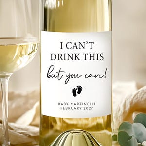 May include: A bottle of white wine with a custom label that reads "I can't drink this but you can!" with baby footprints and the name "Baby Martinelli February 2027". A wine glass is next to the bottle.