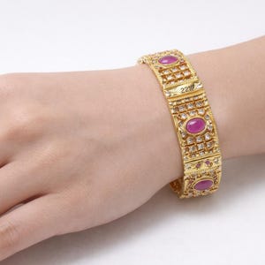 Peut inclure: Un bracelet doré avec un motif répétitif d'éléments rectangulaires et ovales. Le bracelet comporte de petites pierres claires et de plus grandes pierres précieuses roses ovales. Le chiffre "22" est visible.