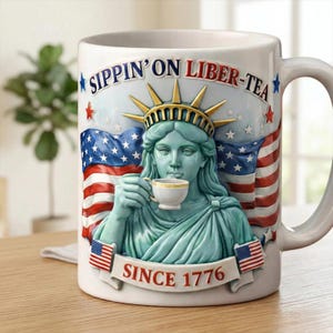 May include: White ceramic mug featuring a sculpted image of the Statue of Liberty holding a teacup. The mug has the text "SIPPIN' ON LIBER-TEA" and "SINCE 1776" with American flags and stars. The mug has a white handle.