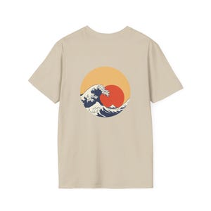 May include: A beige t-shirt with a circular design on the back. The design features a wave, a red sun, and an orange sky. The wave is in blue and white. The t-shirt is a neutral colour.