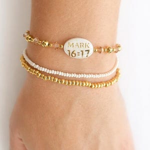 Puede incluir: Tres pulseras apiladas. La pulsera superior tiene cuentas doradas y cristales transparentes, con una cuenta ovalada blanca que dice "MARK 16:17". La pulsera del medio es blanca y la pulsera inferior es dorada.