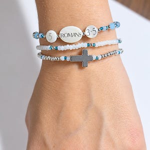 May include: A set of three beaded bracelets featuring white, silver, and blue accents. The top bracelet includes white circular beads with the text ROMANS 5:3-5. The middle bracelet consists of small white beads, and the bottom bracelet features a silver cross charm.