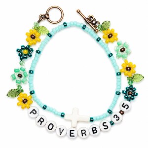 以下が含まれることがあります： 水色のシードビーズ、白い十字架のチャーム、PROVERBS 3:5と綴られた白い文字ビーズを使用した二連のビーズブレスレット。黄色と緑色の花型ビーズ、緑色の葉のアクセント、銅色のトグルクラスプが特徴です。