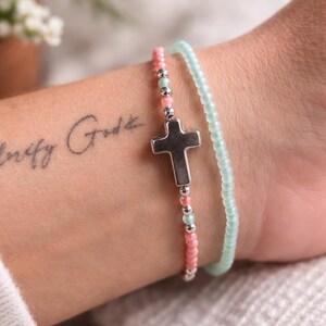 May include: Two bracelets are shown. One is a beaded bracelet with pink and silver beads and a silver cross charm. The other is a light blue beaded bracelet. The bracelets are on a wrist with a tattoo that says "Infinity God".