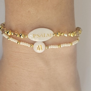 May include: Two beaded bracelets. The top bracelet has a white oval bead with the word "PSALM" in gold lettering. The bottom bracelet has a white round bead with the number "41" in gold lettering. Both bracelets have gold and clear beads.