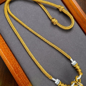 Pode incluir: Um colar dourado com um design de corda trançada. O colar tem um nó na parte superior e detalhes prateados perto do fecho. O colar é exibido em uma superfície cinza dentro de uma moldura de madeira.