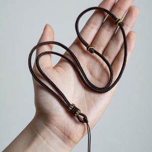Può includere: Una collana in cordino marrone scuro caratterizzata da una trama intrecciata e nodi scorrevoli regolabili. I nodi sono impreziositi da dettagli in filo metallico dorato, offrendo una vestibilità sicura e flessibile per questo gioiello semplice.