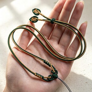 May include: A decorative cord strap featuring a braided design in dark green and metallic gold threads. The strap includes a complex knot detail at the base and two small, circular, braided end pieces, finished with a thin grey attachment loop.