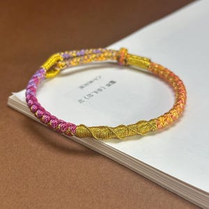Puede incluir: Una pulsera hecha a mano con un diseño tejido. La pulsera presenta un degradado de colores, pasando del rosa al amarillo. Una sección dorada está tejida en el centro. La pulsera está sobre una superficie blanca.