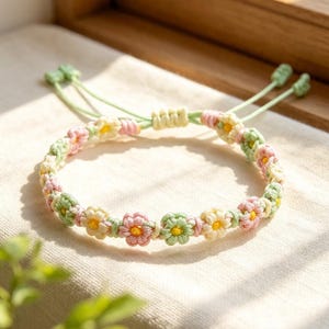 Puede incluir: Una pulsera hecha a mano con un patrón repetitivo de pequeñas flores tejidas a ganchillo en tonos pastel rosa, verde claro y crema. Cada flor tiene un centro amarillo brillante. La pulsera incluye cordones ajustables de color verde claro.
