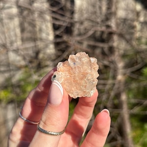 May include: A small, translucent, peach-coloured mineral cluster featuring multiple jagged, crystalline points. The specimen displays a light orange hue with subtle variations in clarity and texture across its multifaceted surface structure.