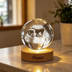 May include: A clear glass sphere featuring a laser-etched 3D ultrasound image of a fetus inside. The sphere rests on a round, light-coloured wooden base that illuminates the glass from below. The name Emma is engraved on the front of the wooden base.