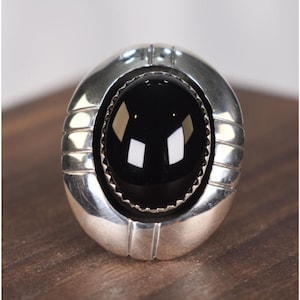 May include: Oval-shaped silver ring with a large, polished black stone center. The ring has a textured, ridged design on the sides and a decorative border around the stone. The ring is set against a dark wooden surface.