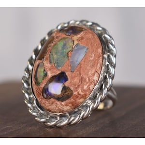 May include: Oval-shaped ring featuring a large, composite stone with a reddish-brown base and colorful opal fragments. The stone is set in a silver-toned, twisted rope-style bezel. The ring's band is also silver-toned.