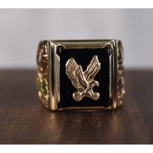 May include: A gold-toned ring with a black square center featuring a detailed gold eagle emblem. The ring has decorative side accents and a polished finish, showcasing a classic design.