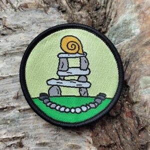 Pode incluir: Remendo bordado redondo com um caracol de desenho animado em cima de uma estrutura de pedra, com uma borda verde e preta. O caracol tem uma concha amarela. O fundo é verde claro com uma base verde e um desenho de corrente.
