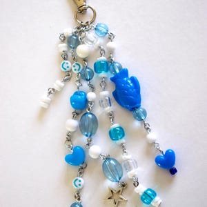 May include: A keychain featuring multiple silver-toned chains adorned with blue and white beads. The beads include various shapes such as hearts, smiley faces, a fish, and a star charm, all attached to a silver-toned lobster clasp.