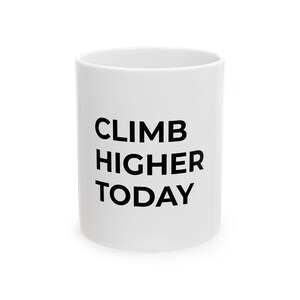 May include: White ceramic mug with the words "CLIMB HIGHER TODAY" printed in bold black letters. The mug has a cylindrical shape and a simple, clean design, suitable for coffee or tea.