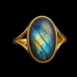May include: A gold ring featuring a large, oval-shaped labradorite gemstone. The stone displays iridescent flashes of blue and yellow across its surface. The metal band has a smooth, polished finish and splits into two sections at the setting.