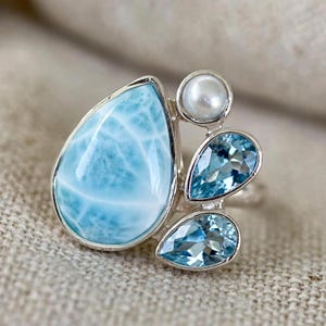 May include: A silver ring featuring a large, pear-shaped, light blue Larimar stone, a small round white pearl, and two smaller, pear-shaped, faceted light blue topaz stones arranged in a cluster on a polished silver band setting.