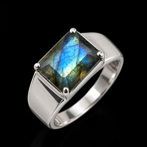 May include: A silver-toned ring featuring a rectangular, faceted labradorite gemstone set in a four-prong mounting. The stone displays iridescent blue and gold flashes across its surface, while the band has a smooth, polished metallic finish.