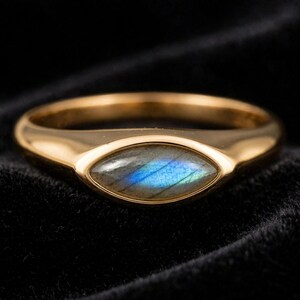 May include: A gold-toned ring featuring a marquise-cut labradorite gemstone set in the center. The stone displays a distinct blue iridescent flash across its surface, while the band has a smooth, polished finish and a tapered design.