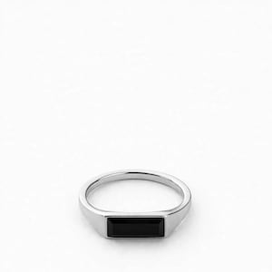 May include: A silver-toned ring featuring a horizontal, rectangular black stone set into the center of the band. The metal band is smooth and polished, creating a clean, continuous loop that holds the dark, flat-faced gemstone securely.